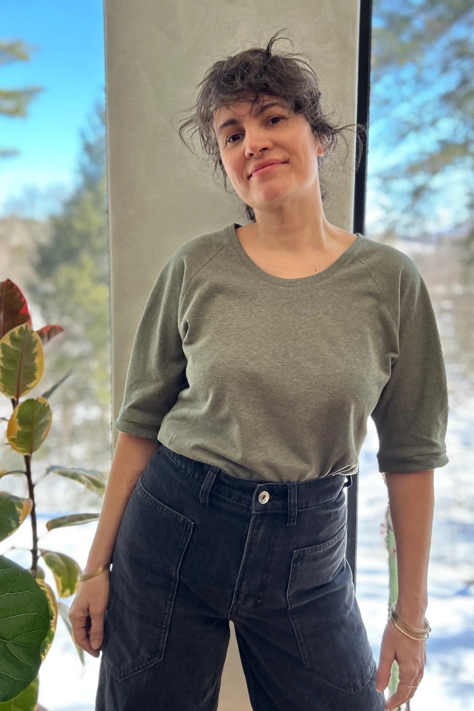 A woman wearing the Vernus Top by Kazak in Sage with elbow-length raglan sleeves with a pleat detail at the shoulders, a round neck, and a fit that tapers at the waist. She is wearing it with dark jeans standing indoors by a window.