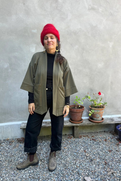 A woman wearing the Cydonia Cardigan by Kazak in Olive, a hip-length open cardigan with 3/4 kimono sleeves and large patch pockets. She is wearing it with black pants and a black top and standing outside.