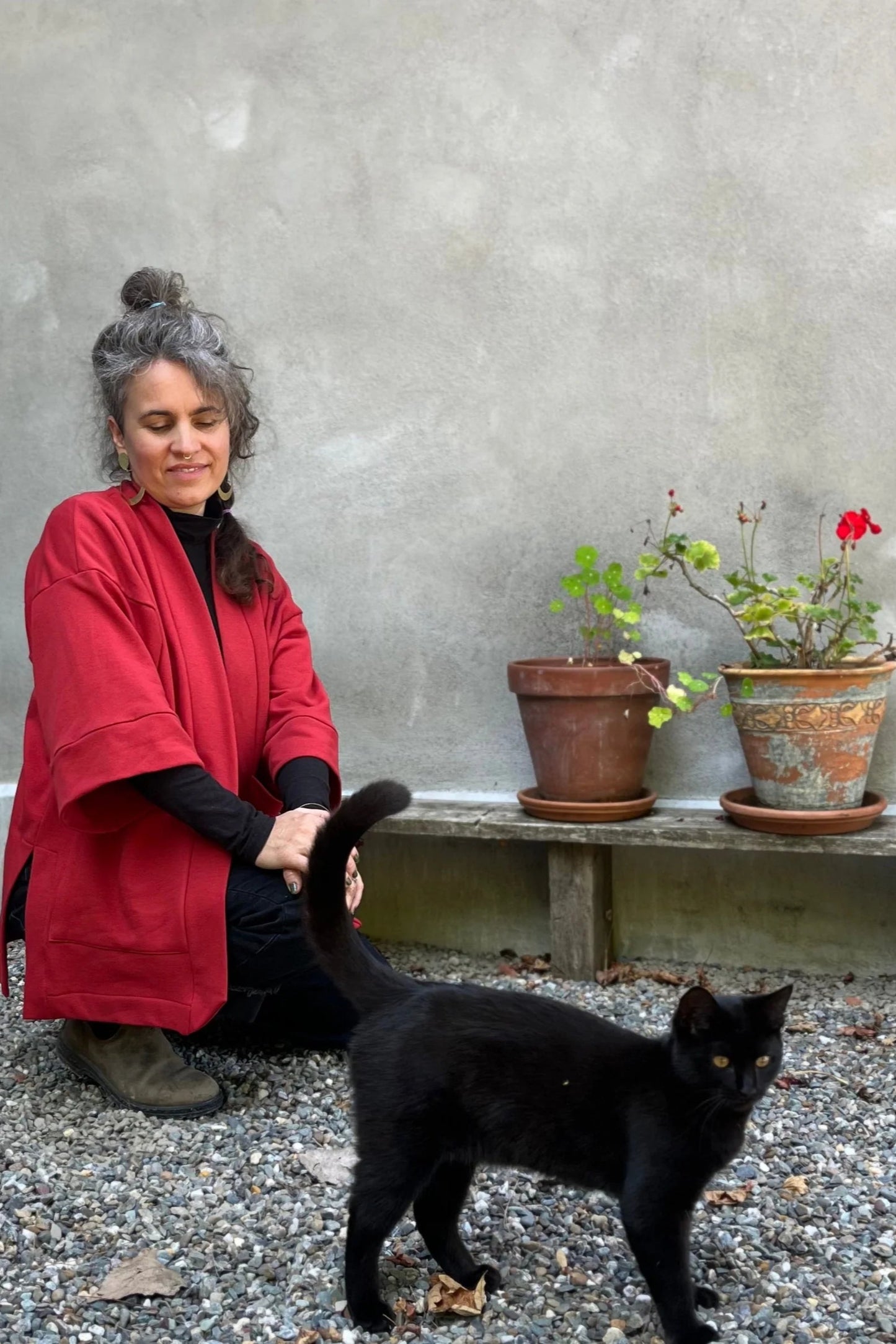 A woman wearing the Cydonia Cardigan by Kazak in Brick, a hip-length open cardigan with 3/4 kimono sleeves and large patch pockets. She is wearing it with black pants and a black top and kneeling outside with a black cat. 