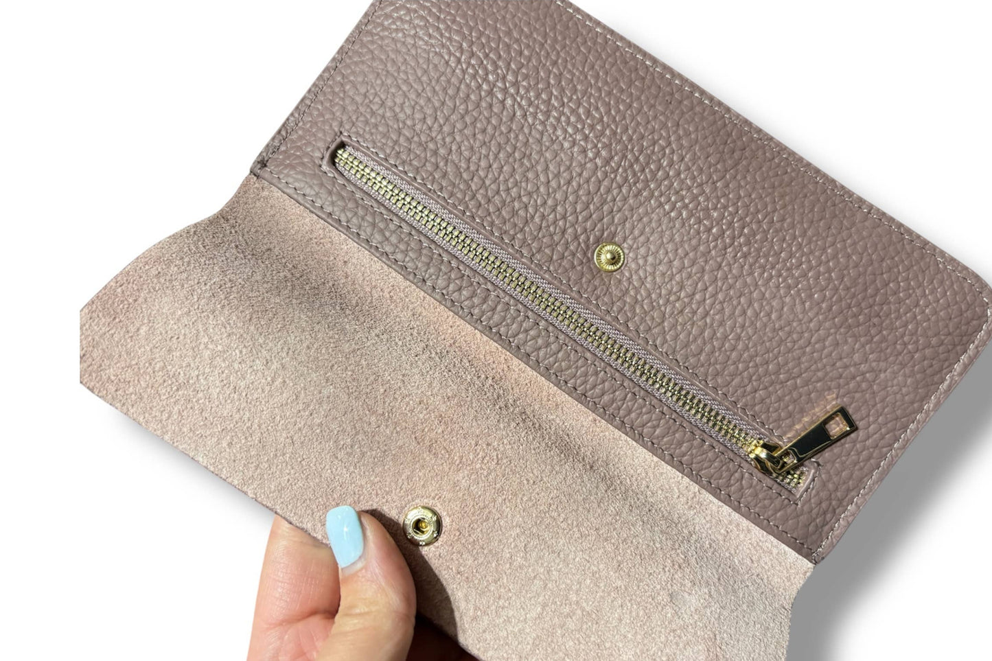 A close-up of a taupe pebbled leather wallet from Armoir being held open by a hand with light blue nail polish, showing a suede-like interior lining and a gold zipper.