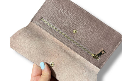 A close-up of a taupe pebbled leather wallet from Armoir being held open by a hand with light blue nail polish, showing a suede-like interior lining and a gold zipper.