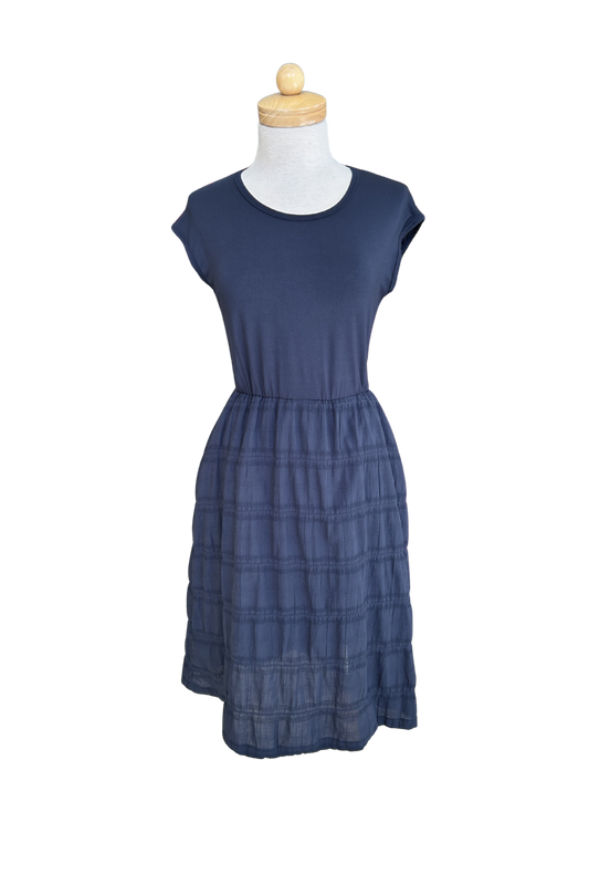 The Mona Dress by Tangente in Navy, a below the knee length dress featuring a bamboo rayon bodice and seersucker skirt, with short sleeves and a round neck, is shown on a mannequin against a white background. 