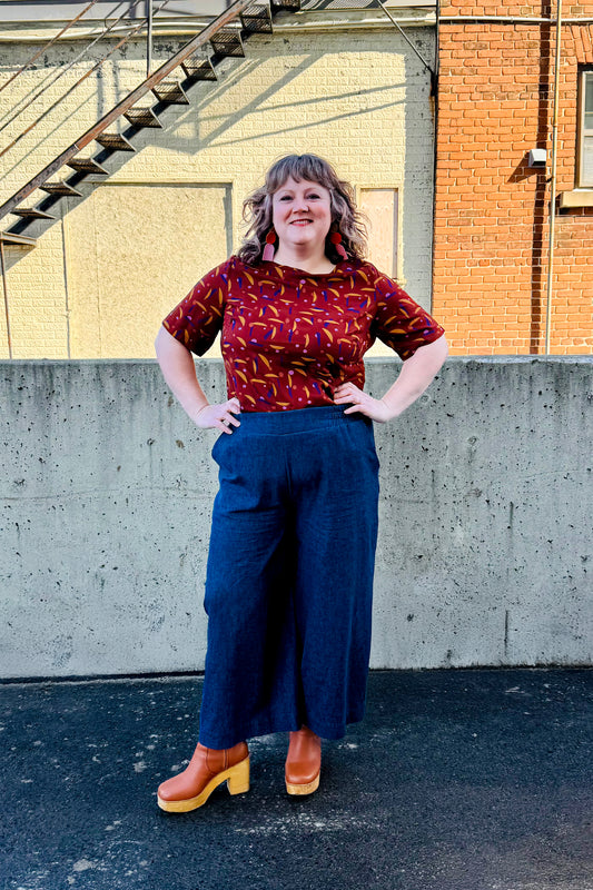 A woman wearing the Mateo Pants by Melow in Denim, ¾ wide leg pants with pull-on waistband that’s elasticized at the back. She is wearing them with the Cecil Box Blouse by Mandala and standing outside in front of a brick building. 