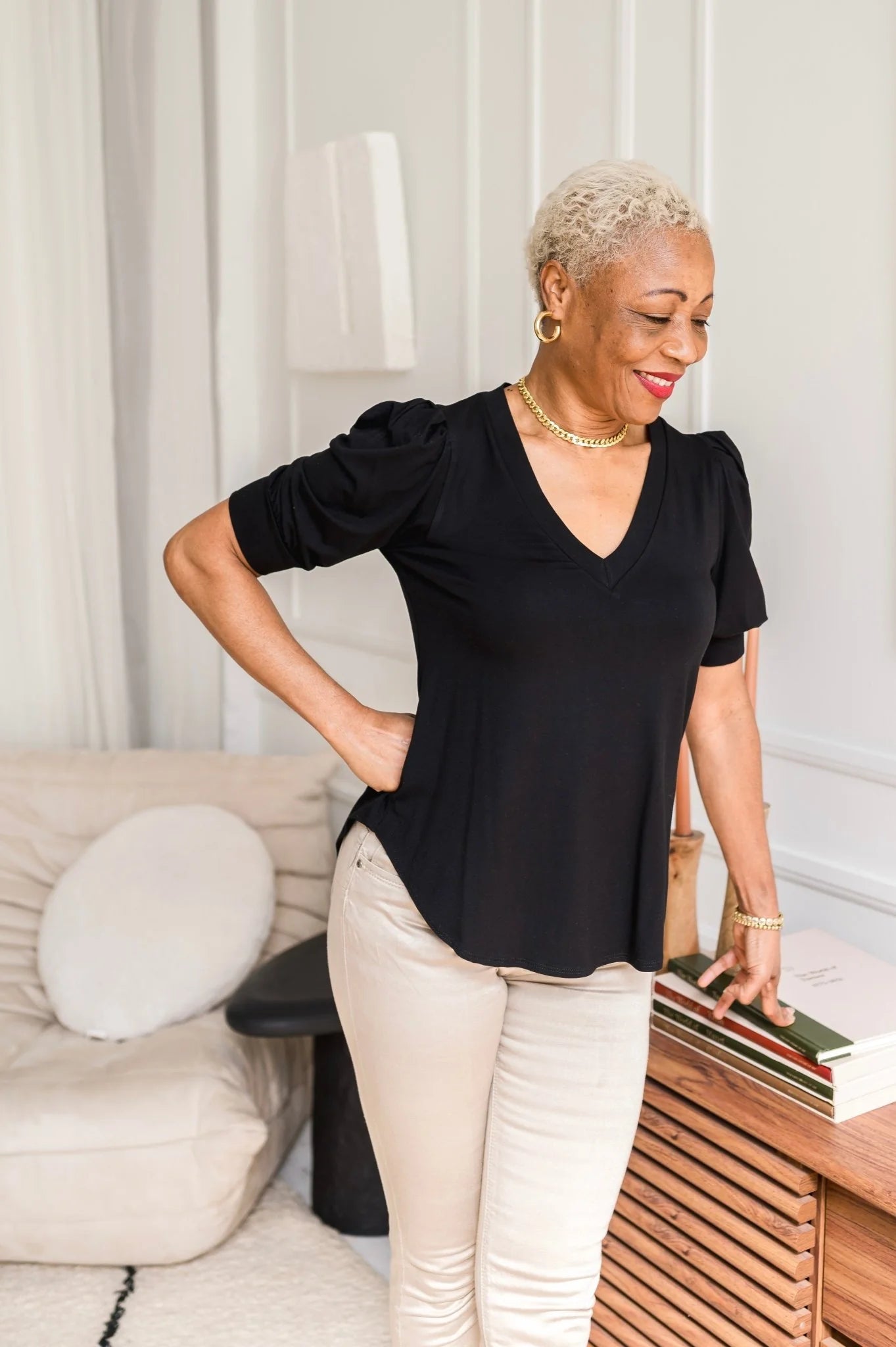 A woman wearing the Darling Tee by Dotty in Black with a v-neck, pleats at the shoulders, elbow-length sleeves, and a rounded hem. She is wearing it with  white jeans and is standing in front of a white wall. 