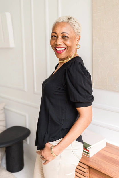 A woman wearing the Darling Tee by Dotty in Black with a v-neck, pleats at the shoulders, elbow-length sleeves, and a rounded hem. She is wearing it with  white jeans and is standing in front of a white wall. 