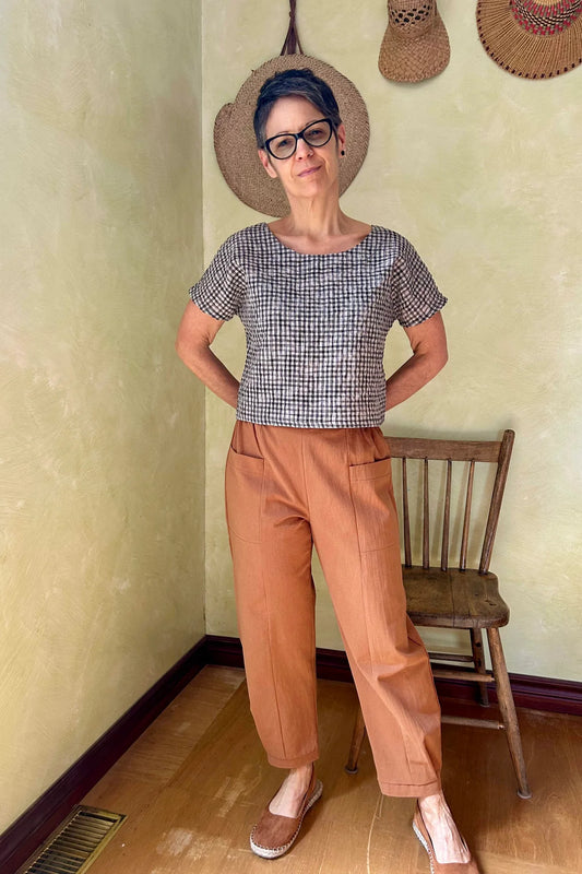 A woman wearing the Brendola Top from Kazak in Black and Cream Vichy/Gingham, a slightly cropped short sleeved top with three brass snaps that can be worn at the back or the front. She is wearing it with the Oxalis Pants in Ginger and standing in a room with a wooden chair. 
