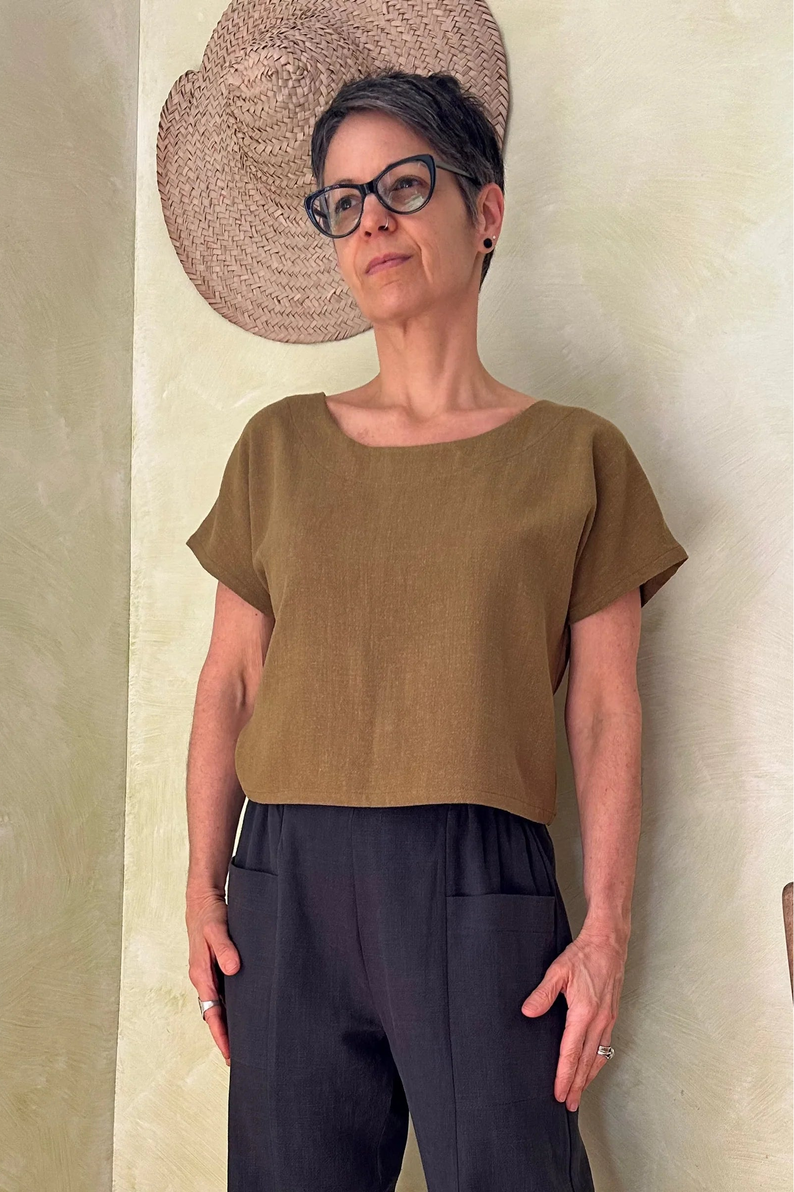 A woman wearing the Brendola Top from Kazak in Mousse, a slightly cropped short sleeved top with three brass snaps that can be worn at the back or the front. She is wearing it with the navy pants and standing in a room with hats on the wall. 