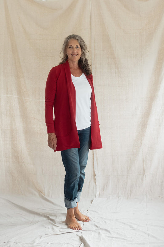 A woman wearing the Briton Cardigan from Blondie in Poppy, a mid-thigh length open cardigan with a small collar and in-seam pockets. She is wearing it with a white tee shirt and jeans and standing in front of a beige background 