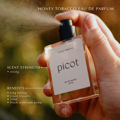 Hand holding a Picot honey tobacco eau de parfum bottle with product details on a blurred natural background
