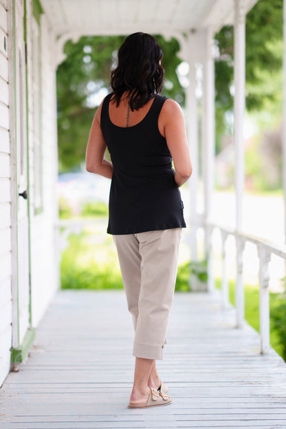 Back view of a woman wearing the Vino 3/4 Pants from Rien ne se Perd in Natural, elastic waisted pants with large pockets, and slim 3/4 legs with side slits with coconut button details. She is wearing them with a black tank and standing on a porch. 