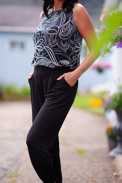 A woman wearing the Pina 7/8 Pants by Rien ne se Perd in Black, with a pull on waist, front pleats, cuffed legs, and a pockets. She is wearing them with a printed black and white top and standing on a porch with greenery in the background. 