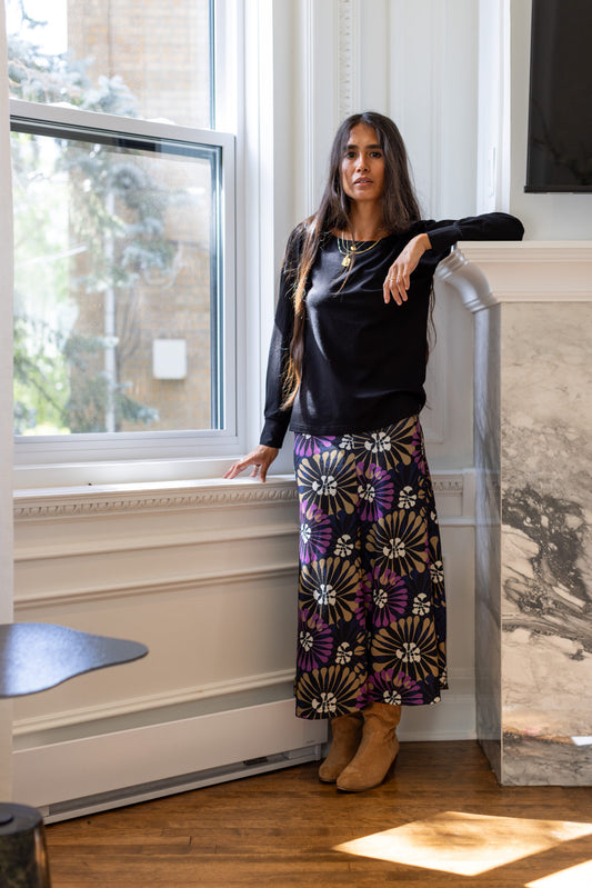 A woman standing in a white living room wearing the Becancour Top by Rien ne se Perd in Black, with a round neck and long slightly puffed sleeves with gathered cuffs. She is wearing it with the Hudson Skirt in Floral.
