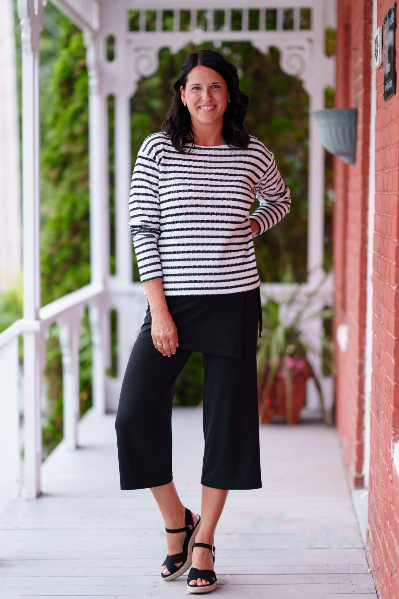 A woman wearing the Collins Reversible Sweater by Rien ne se Perd in Black/White. It's reversible back to front with stripes on side and solid fabric on the other. It has long striped sleeves and a round neck. She is wearing with black pants and standing on a porch. 