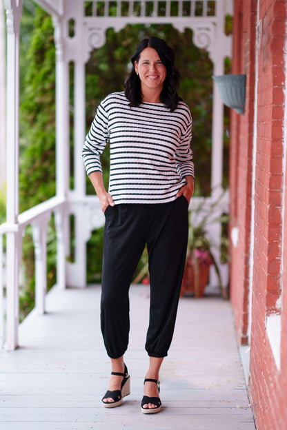 A woman wearing the Collins Reversible Sweater by Rien ne se Perd in Black/White. It's reversible back to front with stripes on side and solid fabric on the other. It has long striped sleeves and a round neck. She is wearing with black pants and standing on a porch. 