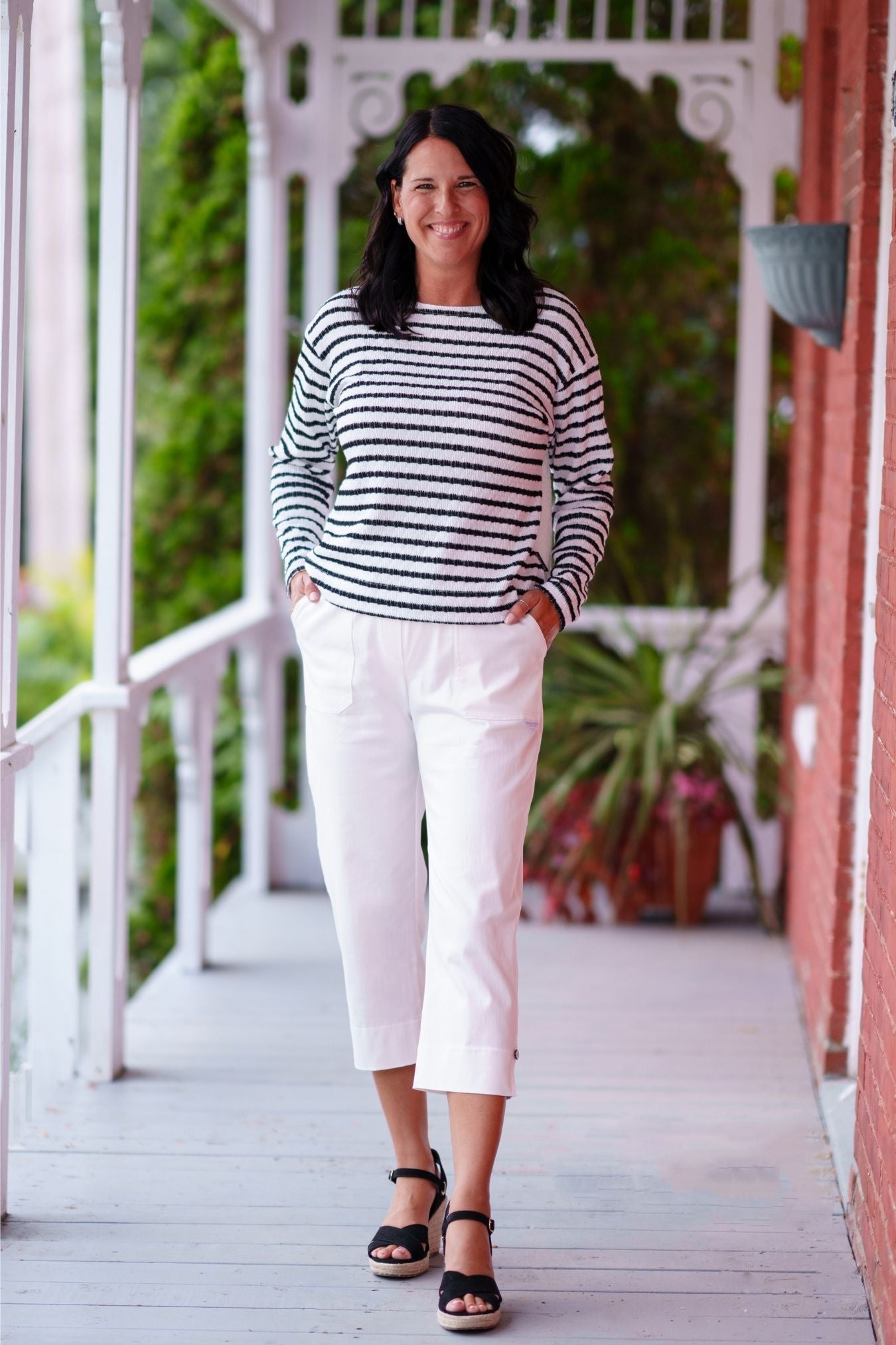 A woman wearing the Collins Reversible Sweater by Rien ne se Perd in Black/White. It's reversible back to front with stripes on side and solid fabric on the other. It has long striped sleeves and a round neck. She is wearing with white pants and standing on a porch. 