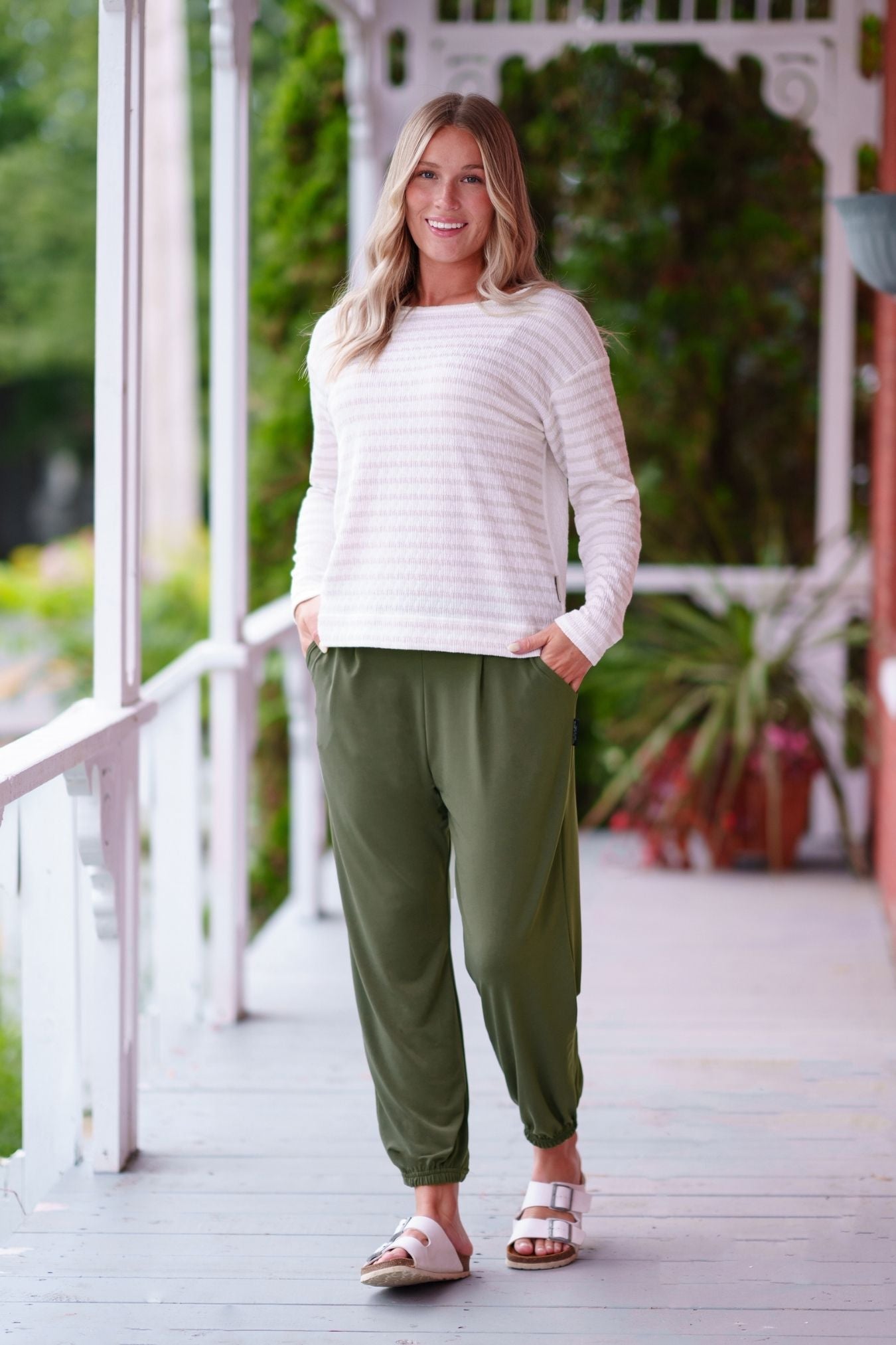 A woman wearing the Pina 7/8 Pants by Rien ne se Perd in Olive, with a pull on waist, front pleats, cuffed legs, and a pockets. She is wearing them with a white sweater and standing on a porch with greenery in the background. 