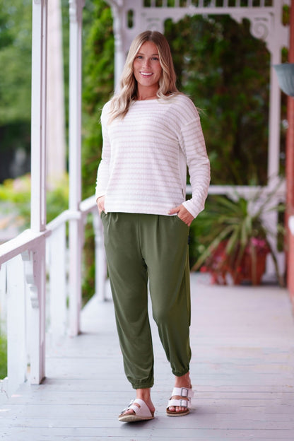 A woman wearing the Pina 7/8 Pants by Rien ne se Perd in Olive, with a pull on waist, front pleats, cuffed legs, and a pockets. She is wearing them with a white sweater and standing on a porch with greenery in the background. 