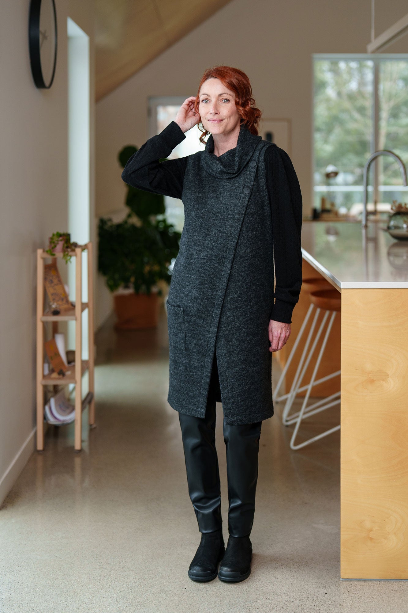 A woman wearing the Resinosa Pants by Rien ne se Perd in Black, slim fitting pants with faux leather at the front and plain fabric at the back, with a wide pull on waistband. She is wearing them with a long charcoal vest and standing in a kitchen.