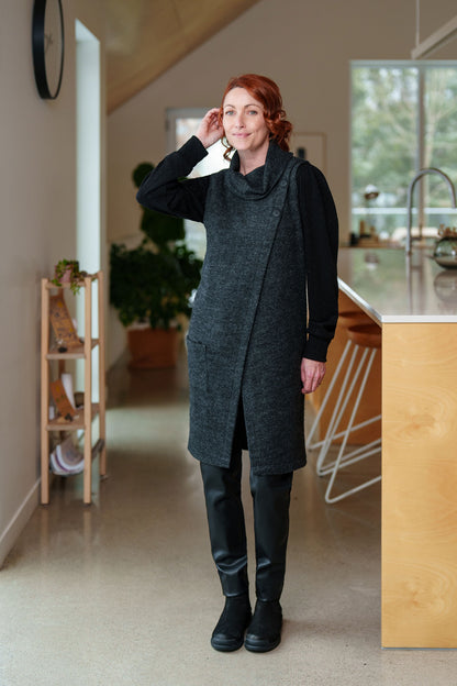A woman wearing the Resinosa Pants by Rien ne se Perd in Black, slim fitting pants with faux leather at the front and plain fabric at the back, with a wide pull on waistband. She is wearing them with a long charcoal vest and standing in a kitchen.