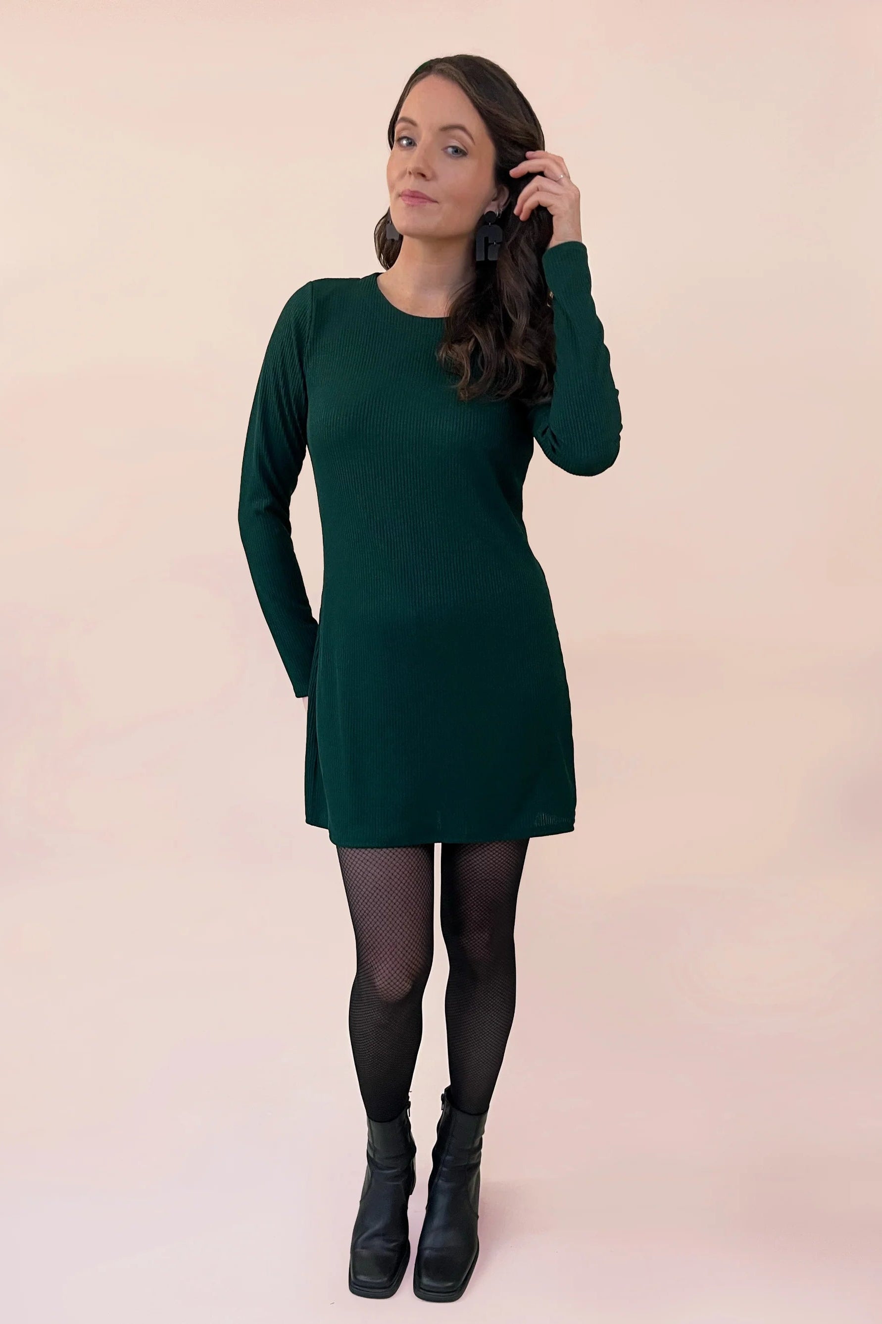 Woman wearing a the Nebulous Dress by Canopée in Green, a mid-thigh length dress with long sleeves in rib knit. She is standing in front of a  beige background. 