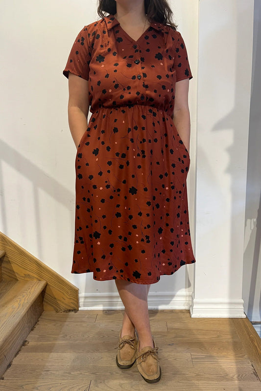 Neck down view of a woman wearing the Paloma Dress by Cherry Bobin in Black Floral with Bronze, with a classic collar, vintage brass buttons, short sleeves, an elastic waist, and a full below-the-knee skirt. She is standing by a staircase. 