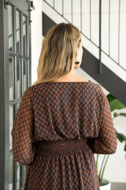 Back view of a woman wearing the Spritz Dress by Cherry Bobin in Brown Pattern, featuring puff sleeves, a v-neck,  cinch waisted, and a full knee-length skirt. She is standing in a white room with a black staircase.
