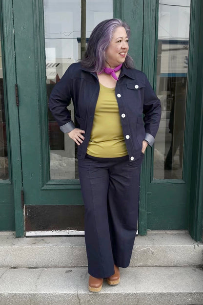 A woman wearing the Bella Knit Jacket by Julei, a denim style jacket with large contrast buttons, a chest pocket and two front patch pockets. She is wearing it with a green tank and the matching Brooke Knit Trousers and standing outside a green door. 