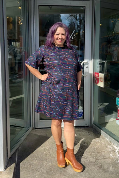 Christina is wearing the Cecily Simple Shift Dress from Mandala in Dark Stix print. It has a high neck, short sleeves, a knee-length hem, French darts, and elastic at the back of the waist. She is standing outside of Flock. 