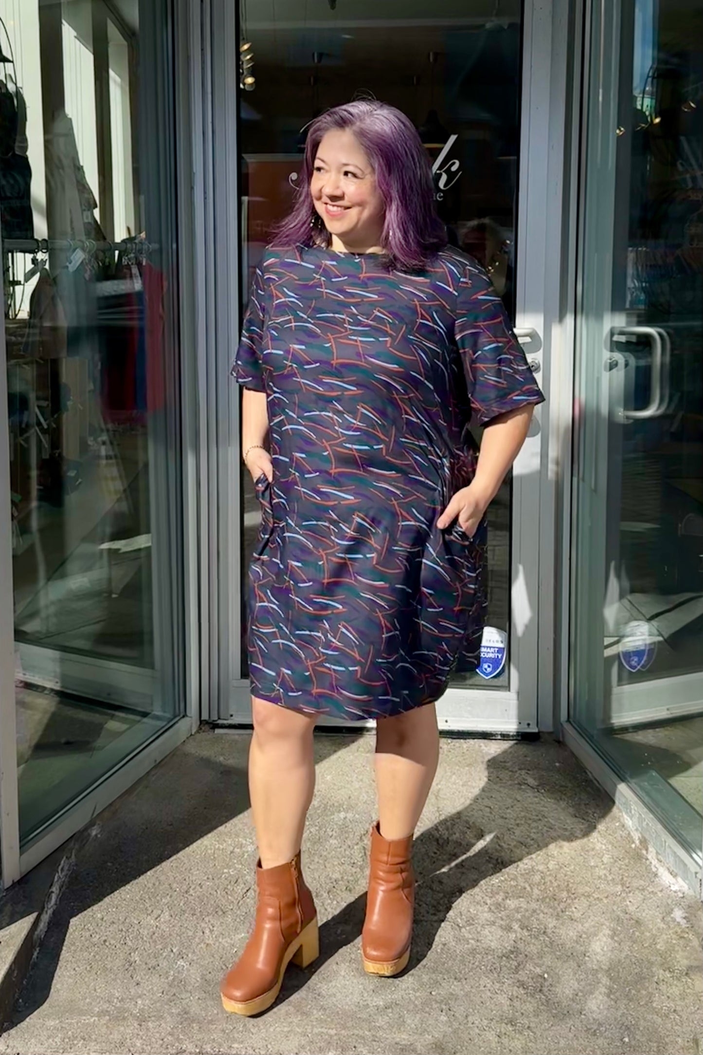 Christina is wearing the Cecily Simple Shift Dress from Mandala in Dark Stix print. It has a high neck, short sleeves, a knee-length hem, French darts, and elastic at the back of the waist. She is standing outside of Flock. 