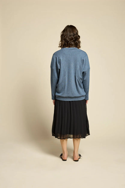 Back view of a woman wearing the Flowers Skirt by Cokluch in Black, a midi-length pleated skirt with a sheer overlayer and an elastic waist. She is wearing it with a blue cardigan and standing in front of a beige background. 