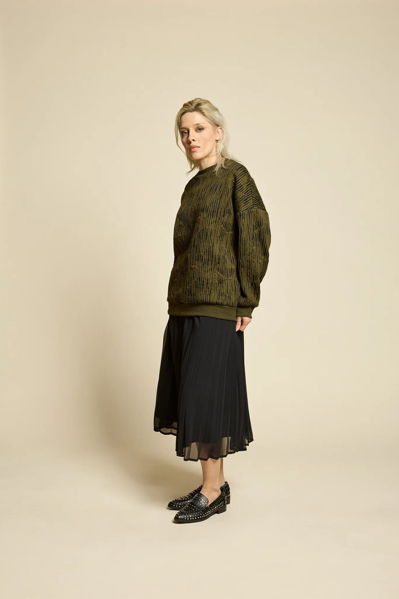 A woman wearing the Flowers Skirt by Cokluch in Black, a midi-length pleated skirt with a sheer overlayer and an elastic waist. She is wearing it with a the Hotel Sweater in Olive  and standing in front of a beige background. 