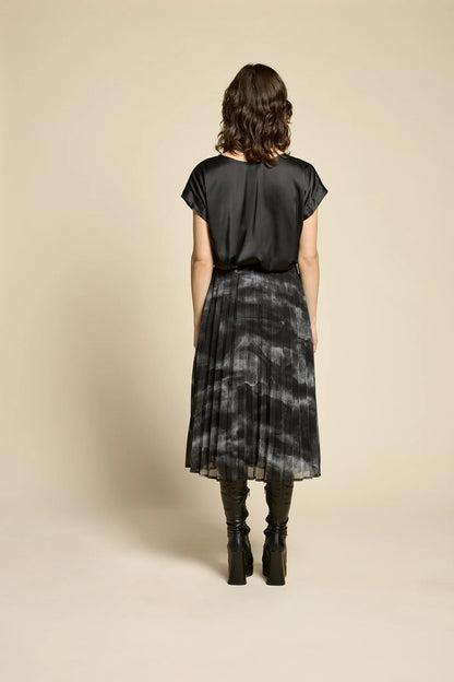 Back view of a woman wearing the Flowers Skirt by Cokluch in Mist Black, a midi-length pleated skirt with a sheer overlayer and an elastic waist. She is wearing it with a black top and standing in front of a beige background. 