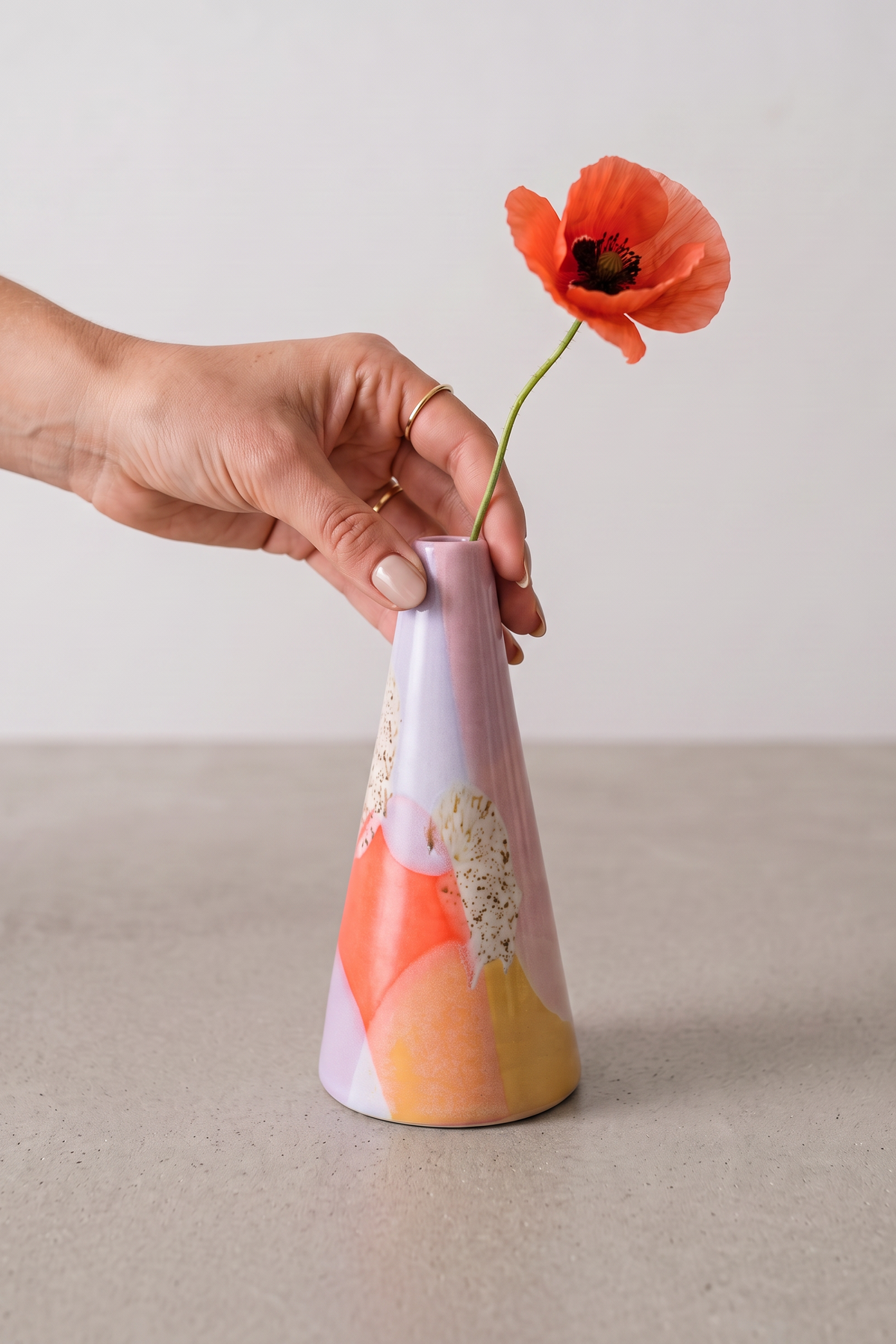 A hand holds a small tapered ceramic vase with a pastel multicolor abstract design and a single orange poppy flower in it, set on a light gray surface against a neutral light gray background.