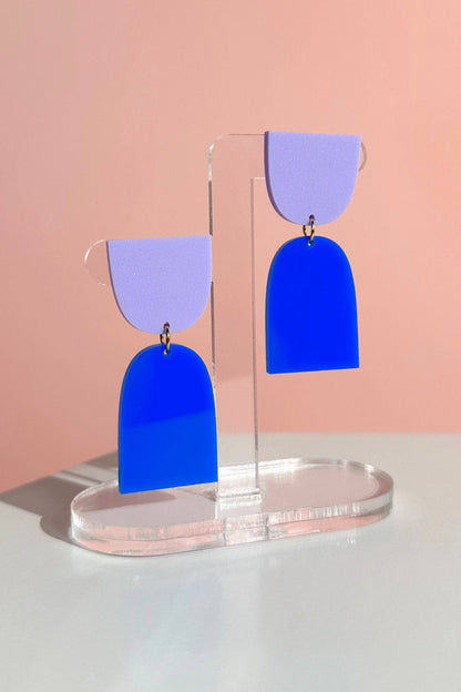 Blue and purple earrings on a clear stand against a pink background