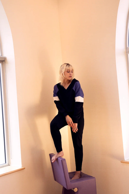 A woman wearing the Sinclair Sweatshirt by Eve Lavoie in Black, a relaxed sweatshirt with a V-neck and colour-blocked panels. She's wearing it with black pants and standing on a purple chair in a beige room. 