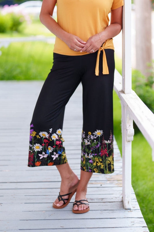 Waist down view of a woman wearing the Amaretto 7/8 Gaucho Pants from Rien ne se Perd in Black with a floral print hem. She is wearing them with a yellow top and standing on a porch. 