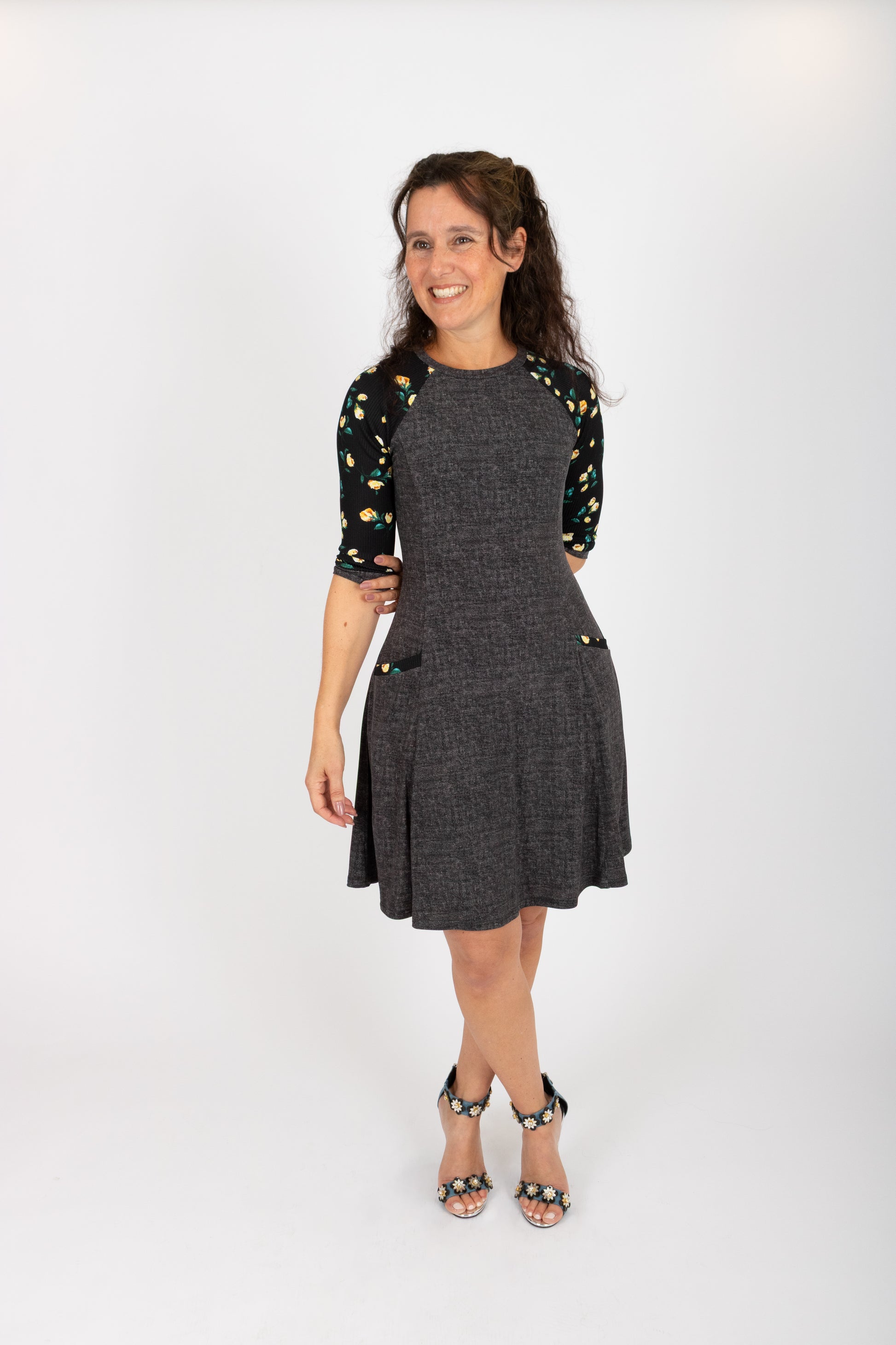 A woman wearing the Raphaelle Dress from Infime in Black, an A-line knee-length dress with 3/4 raglan sleeves that feature a floral print and pockets with a floral detail. She is standing in front of a white background