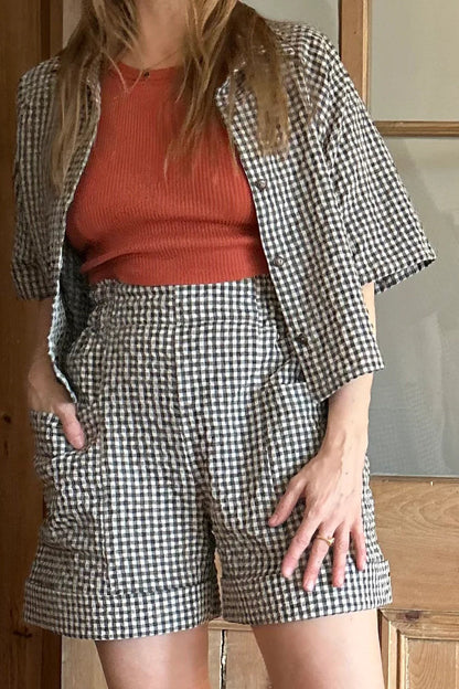 Torso view of a woman wearing the Iris Shorts by Kazak in Teal Vichy/Gingham, with a smooth front waist, elastic at the back, large pockets, and mid-thigh length. She is wearing them with an orange tank and matching open top and standing in a room with a door. 