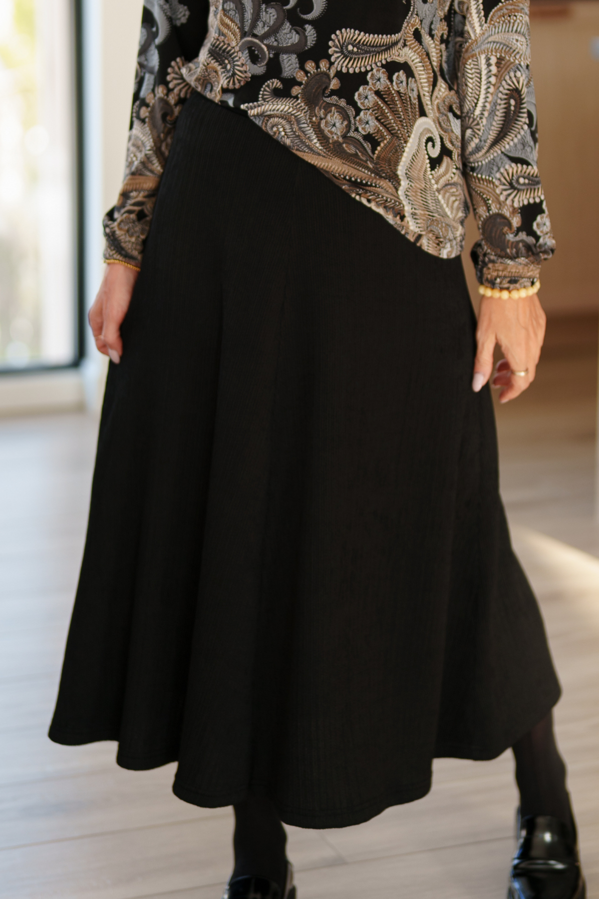 Waist down view of a woman wearing the Memphre Skirt from Rien ne se perd in Black, a long skirt made from a stretchy pleated fabric that gives a corduroy look. She is wearing it with a paisley top and standing in a house.  