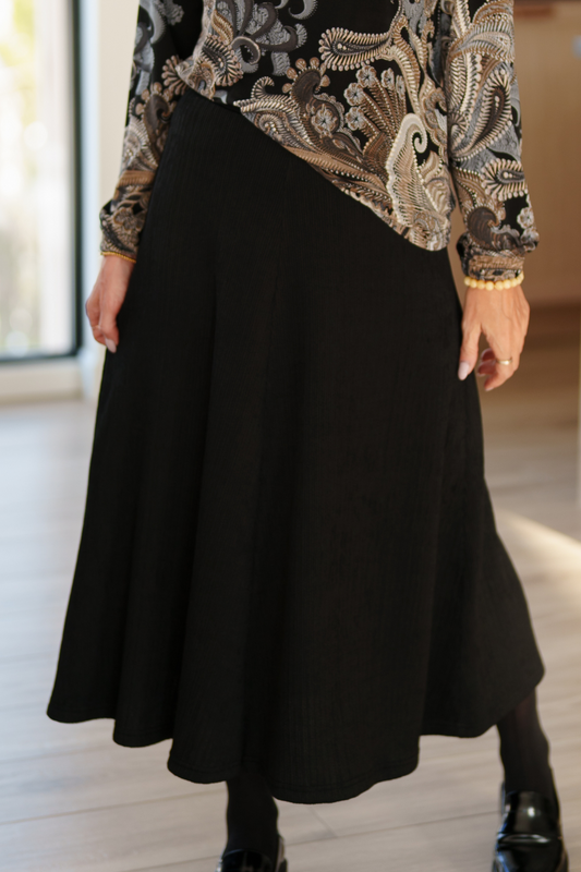 Waist down view of a woman wearing the Memphre Skirt from Rien ne se perd in Black, a long skirt made from a stretchy pleated fabric that gives a corduroy look. She is wearing it with a paisley top and standing in a house.  