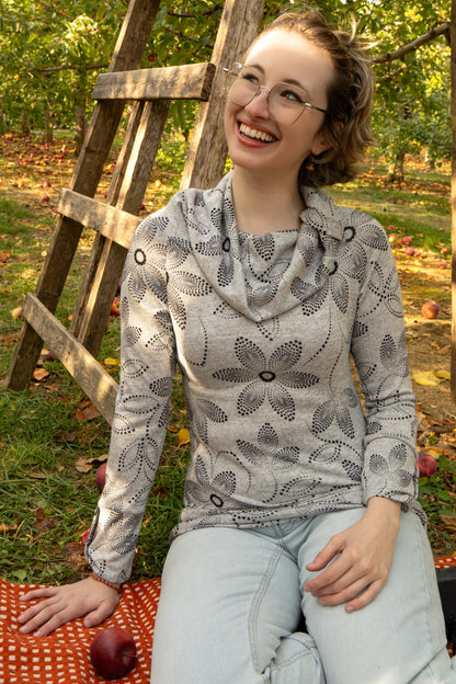 A woman wearing the Teddy Pearl Sweater by Karkass in Grey and Black, a cowl neck sweater with a floral pattern in black dots. She is sitting outdoors with trees and grass in the background.