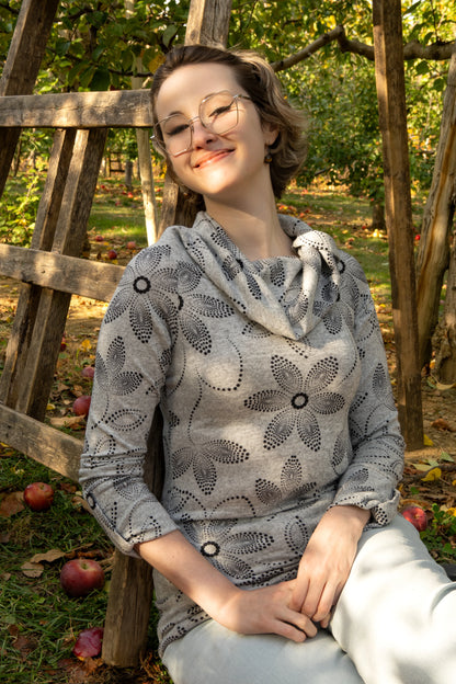 A woman wearing the Teddy Pearl Sweater by Karkass in Grey and Black, a cowl neck sweater with a floral pattern in black dots. She is sitting outdoors with trees and grass in the background.