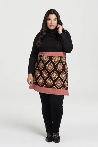 A woman wearing the Ella Pants from Kollontai in Black, slim pants with a wide pull on waistband. She's wearing them with patterned tunic and standing in front of a grey background. 