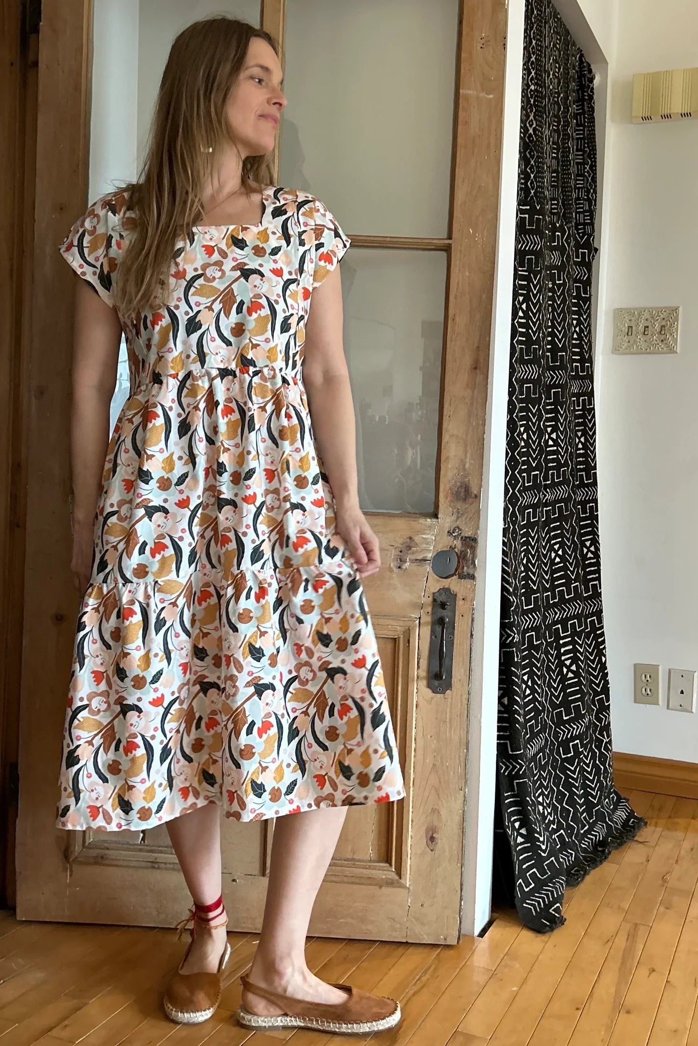 Woman wearing the Lavendula Dress by Kazak in Floral with cream, a short sleeved square neck midi dress with three ruffles. She is standing in a room with wooden floors and a doo
