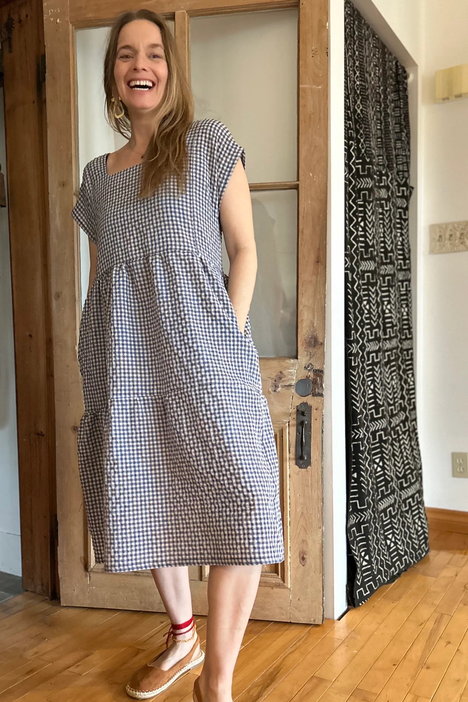 Woman wearing the Lavendula Dress by Kazak in Sky Vichy/Gingham, short sleeved square neck midi dress with three ruffles. She is standing in a room with wooden floors and a door.