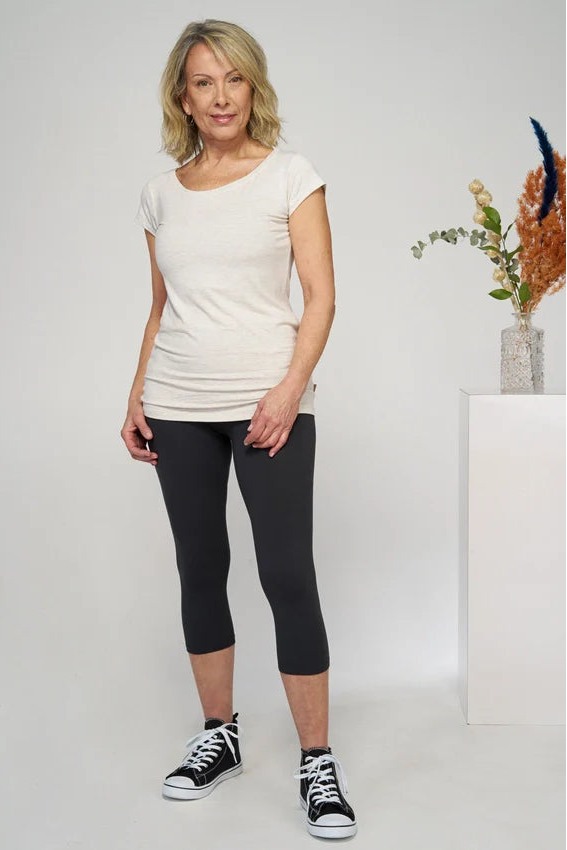 A woman wearing the 3/4 Bojan bamboo leggings from Moovment in Black, with a white t-shirt. She standing against a white background beside a pillar with a plant on it. 