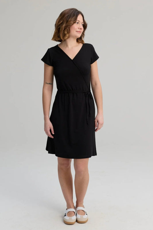 A woman wearing the Libellule Dress by Message Factor in Black, a short sleeved wrap dress that falls to the knee. She is standing against a plain background. 