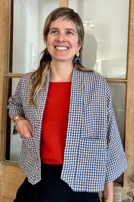 A woman wearing the Magnolia Jacket from Kazak in Sky Vichy/Gingham print, a kimono style jacket with 3/4 sleeves and large patch pockets. She is waering it with a red top and navy skirt and standing in front of a door. 