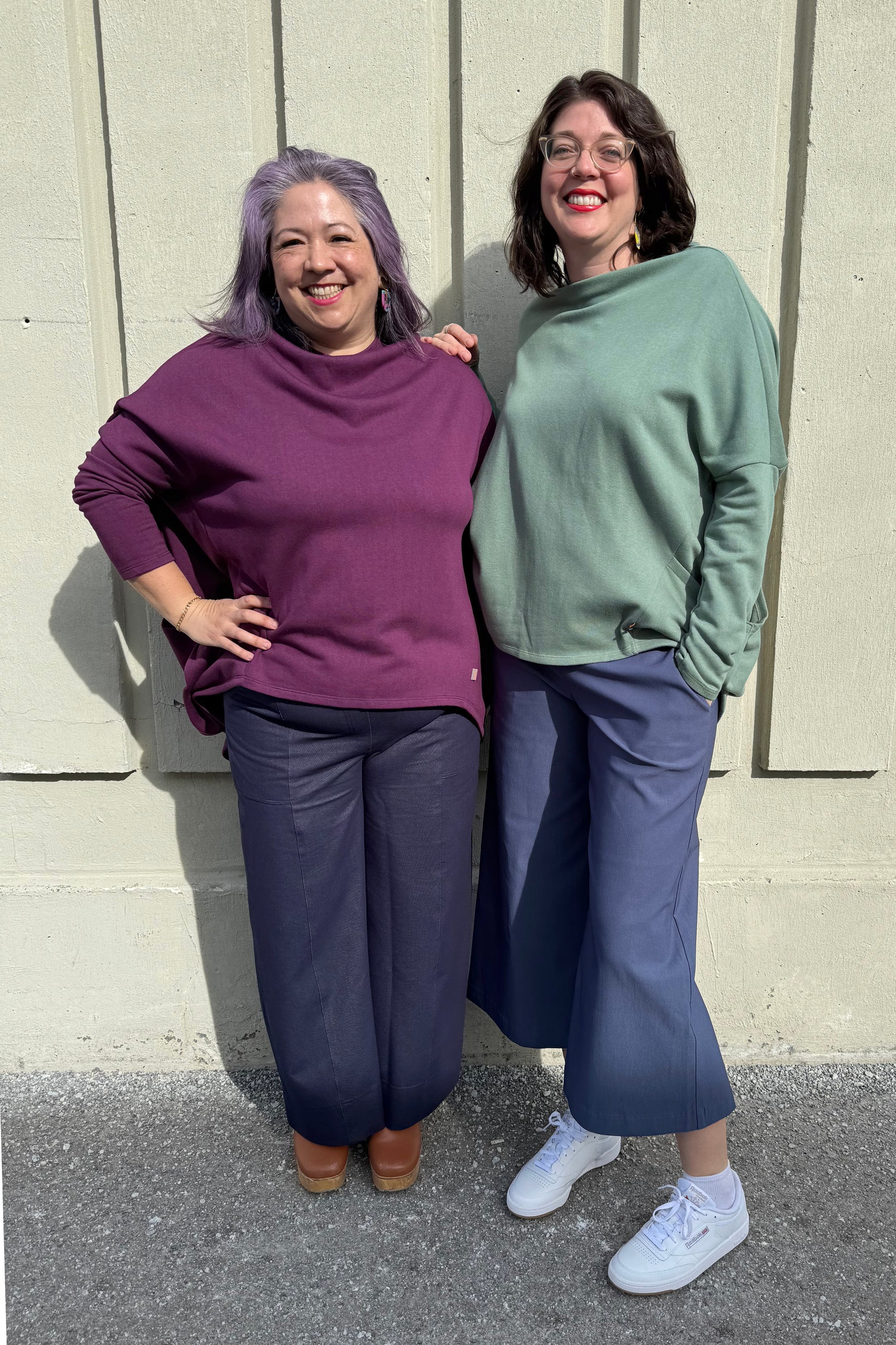 Two women wearing the Meeso Sweater by Moovment, one in Sage and one in Raisin. The top has big side pockets, a wide mock turtleneck, and long dropped sleeves. They are standing together against a beige wall, wearing  blue pants.