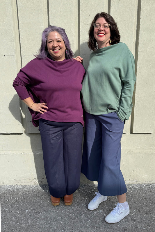 Two women wearing the Meeso Sweater by Moovment, one in Sage and one in Raisin. The top has big side pockets, a wide mock turtleneck, and long dropped sleeves. They are standing together against a beige wall, wearing  blue pants.